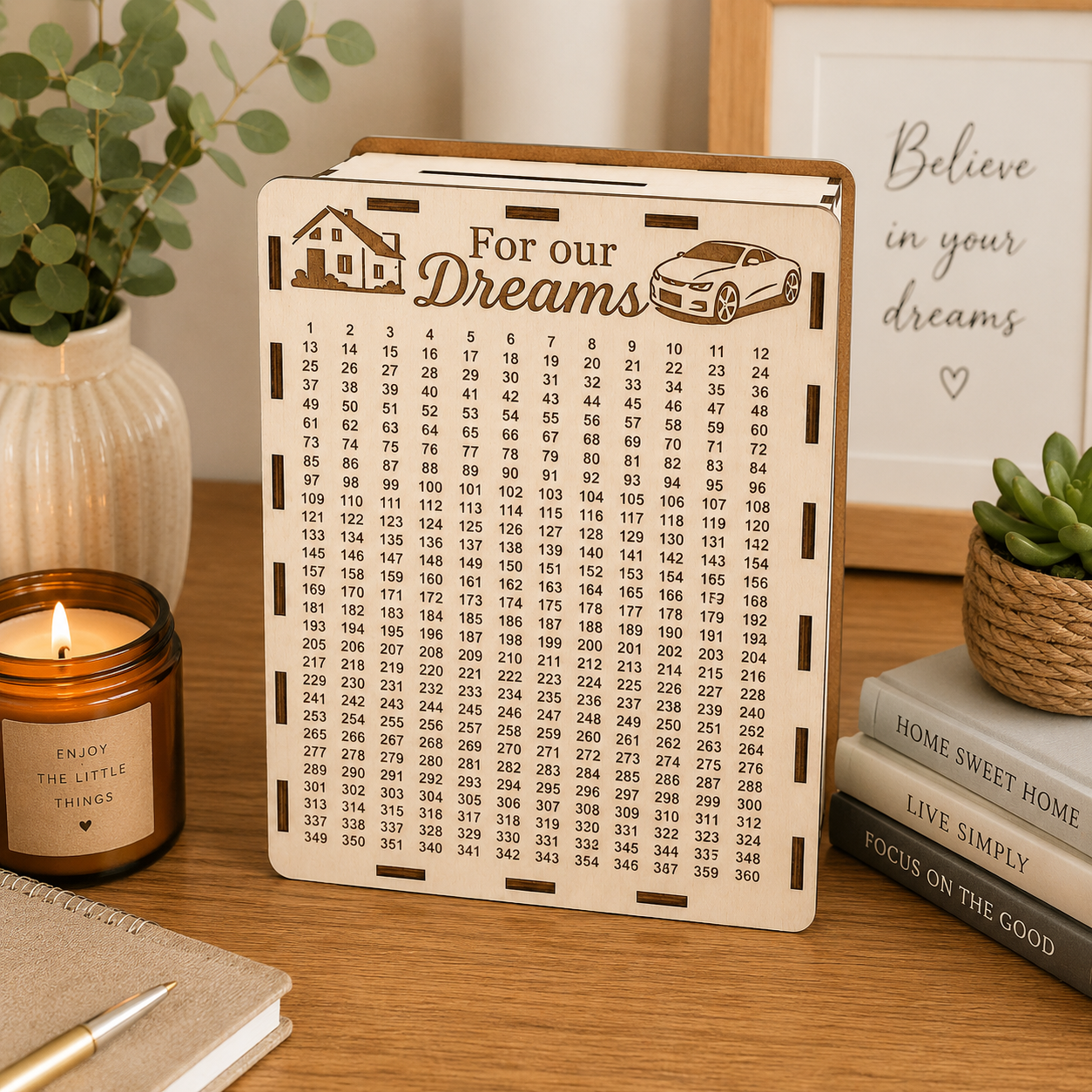 Light wooden “For our Dreams” savings box standing on a wooden table in a warm cozy home interior with a candle, plants, books, and soft natural light.